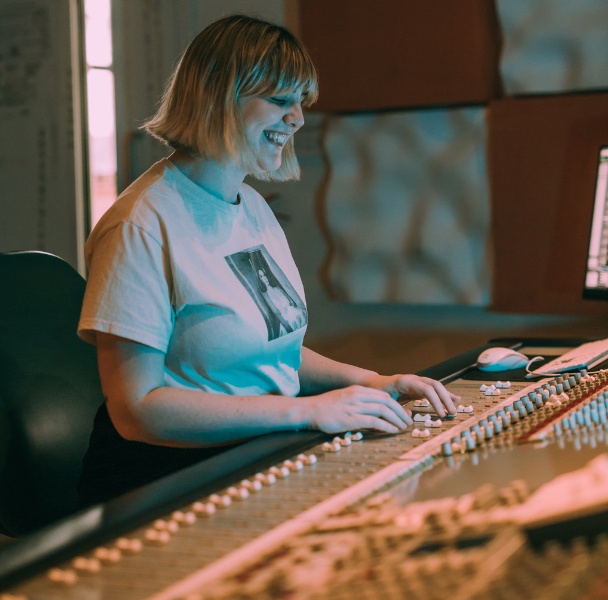 A close-up of a student smiling whilst operating a sound desk in a recording studio.