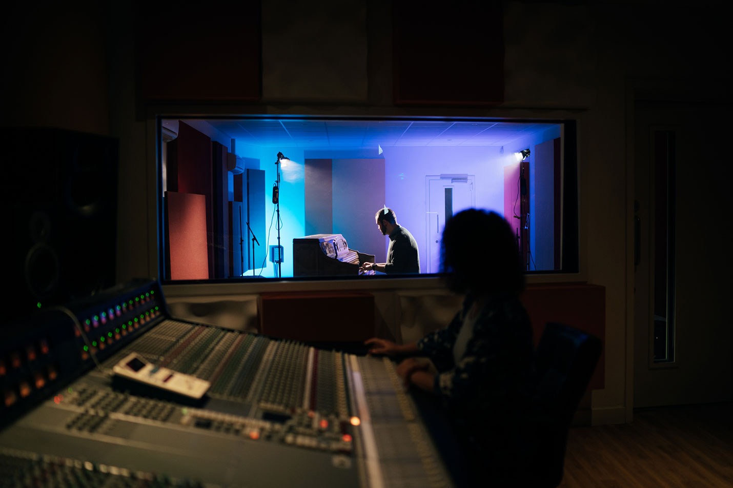 A picture of a pair recording in a studio, one on the production desk, the other playing piano.