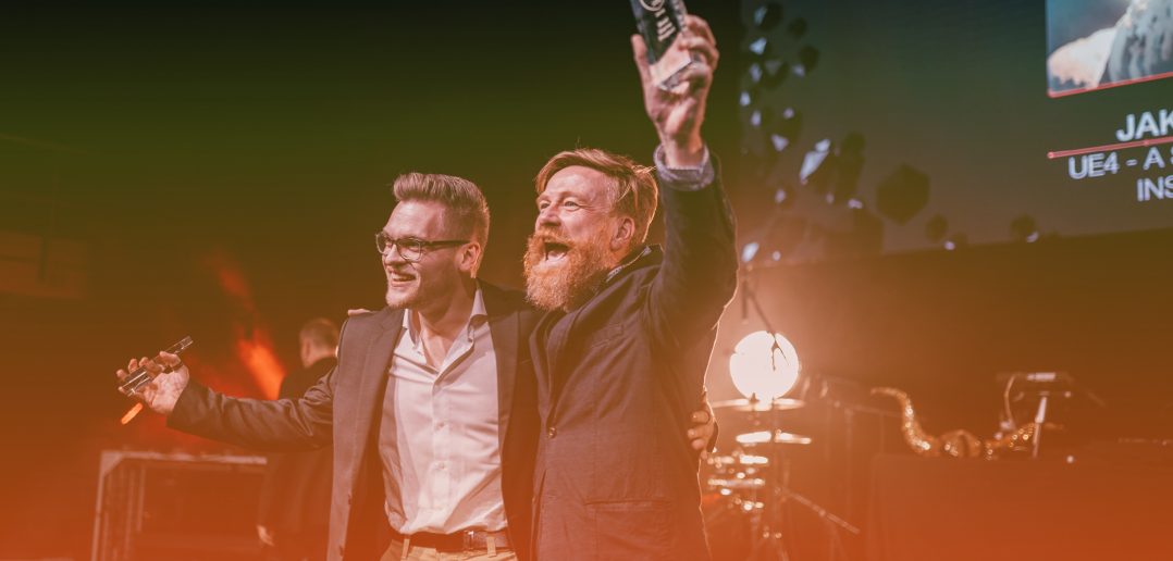 A close-up of a pair of individuals at the SAE convention, holding up their awards and smiling towards the crowd.