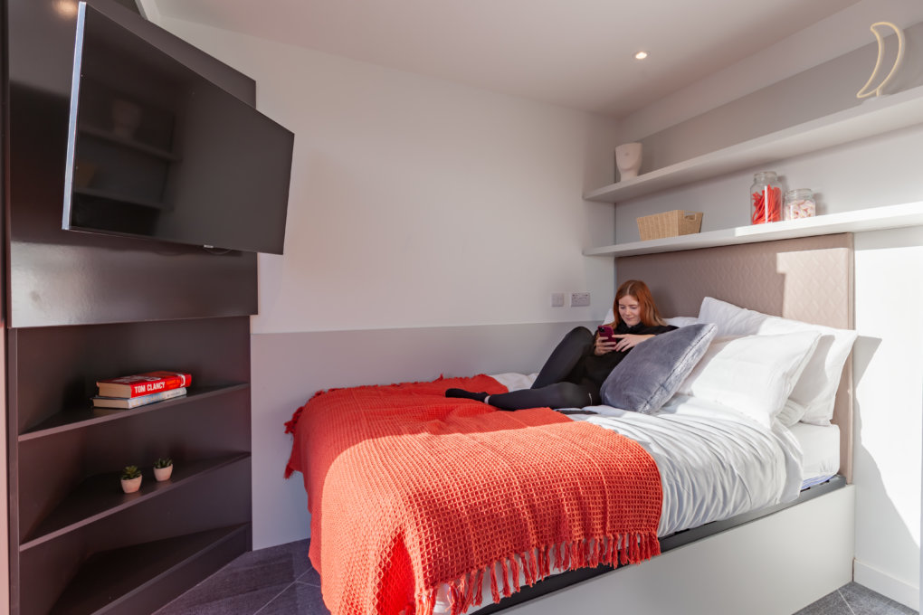 A picture of a student resting on the bed of their student accommodation whilst using their phone.